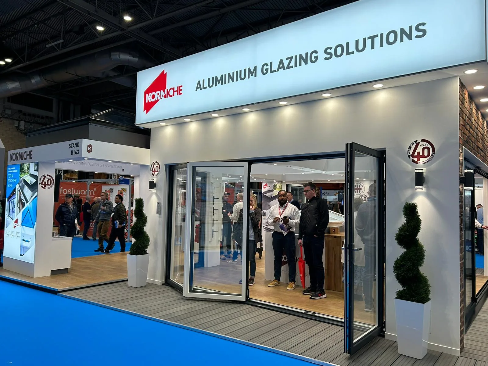 wide shot of the Korniche stand at the National Homebuilding & Renovating Show. Attendees crowd the stand, chatting with the team and examining a roof lantern and bi-folding door on display.