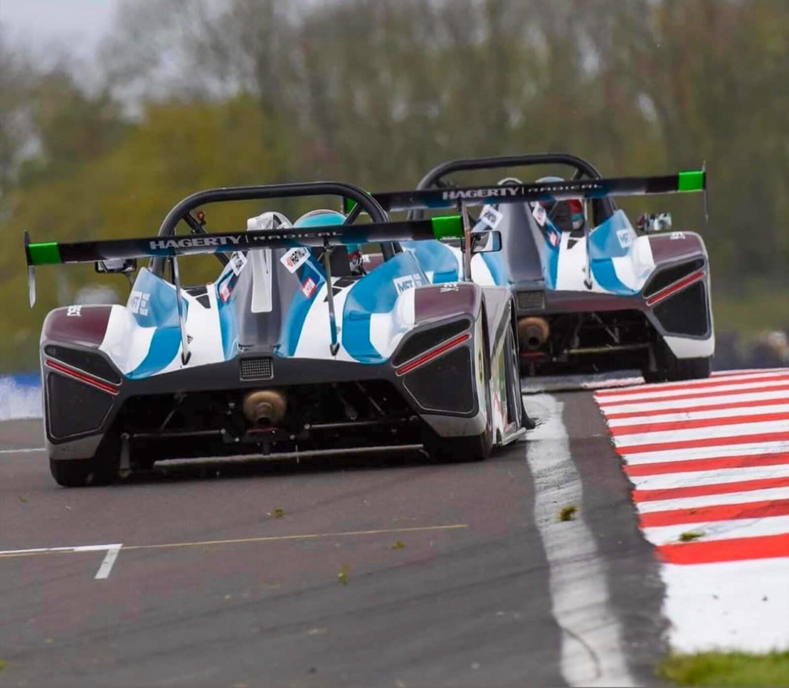 wo North Motorsport Race cars, driven by Bradley Gaunt and Ashley Gaunt, side-by-side rounding a corner with their wheels near the curb.