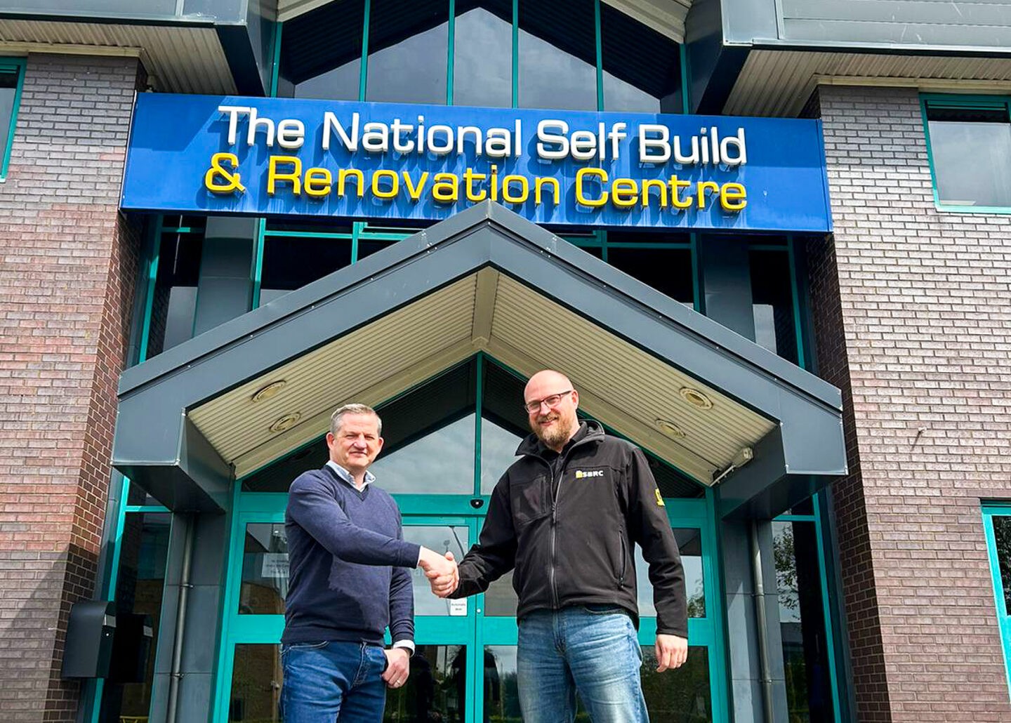 A photo of two men shaking hands in front of a large sign that reads "The National Self Build & Renovation Centre." The man on the left is smiling and wearing a Korniche jacket (Ian Bousfield, Made For Trade's Marketing Manager). The man on the right is smiling and wearing a fleece with the NSBRC logo (Gareth Nicholas, NSBRC Business Development Manager).