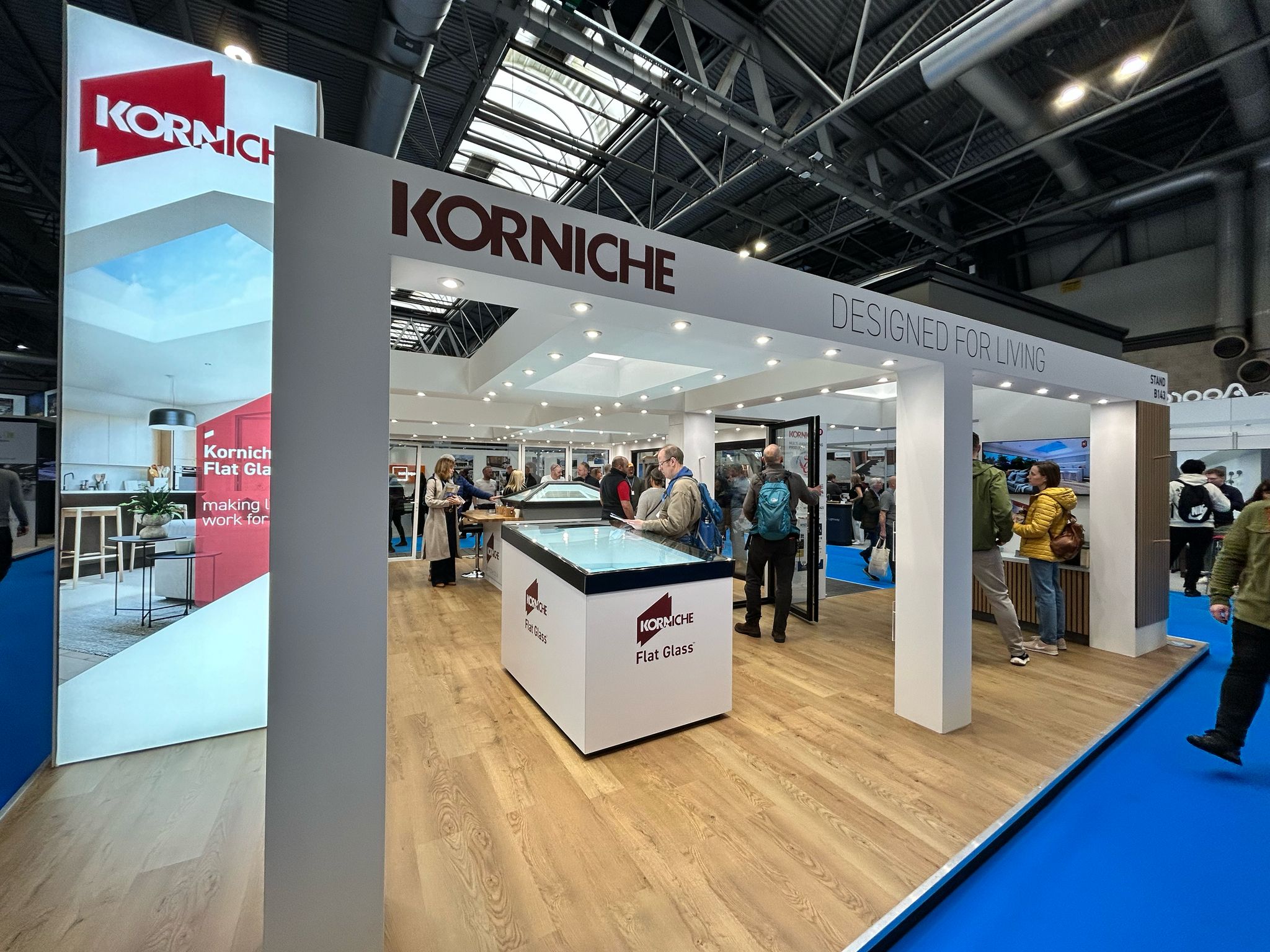 Korniche Stand B143 exterior, "Korniche Designed For Living" signage, showing Flat Glass, Bifold Doors, and Roof Lantern. Visitors interacting with products.