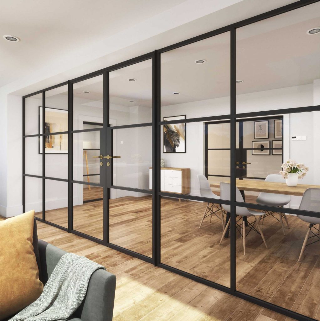 Interior view of a Korniche glass partition system with black frames, featuring double doors and fixed sidelights, separating a living area with a grey sofa and a dining room with a wooden table and chairs. Natural light floods the space through the clear glass panels.