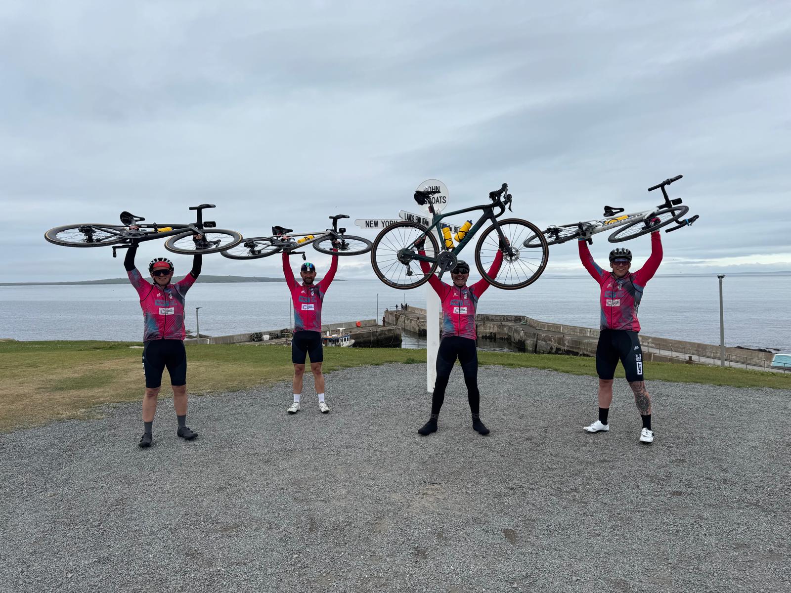 The four Handlebar Heroes cyclists, Gareth Turner, Chris Wann, Leigh Greenwood, and Ashley Parkinson, joyfully hold their bikes triumphantly over their heads at John O'Groats, celebrating the completion of their Lands End to John O'Groats challenge and their fundraising efforts for Cancer Research UK and Andy's Man Club.
