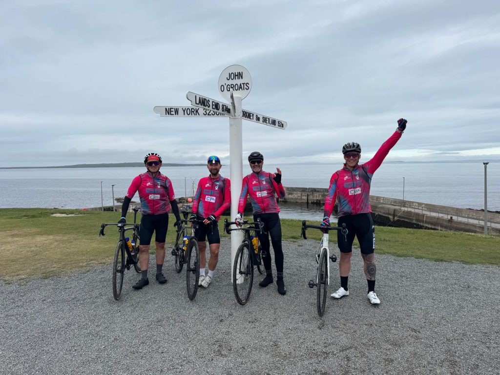 Four cyclists, Gareth Turner, Chris Wann, Leigh Greenwood, and Ashley Parkinson, known as the Handlebar Heroes, stand with their bikes at the iconic John O'Groats signpost, celebrating completing their Lands End to John O'Groats challenge, which raised funds for Cancer Research UK and Andy's Man Club.