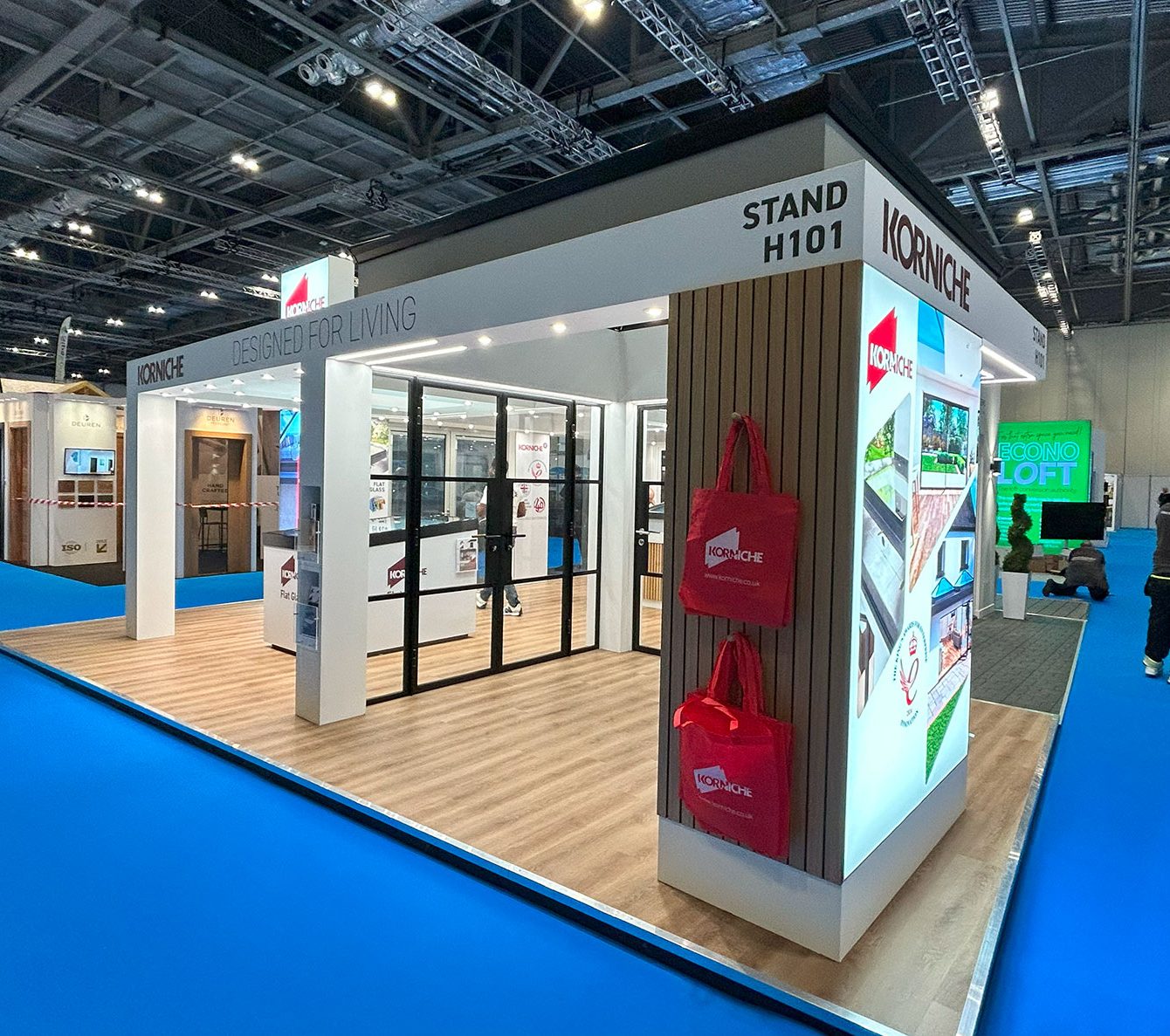The Korniche stand, H101, at a trade show, featuring the "Designed For Living" slogan and red tote bags for visitors.